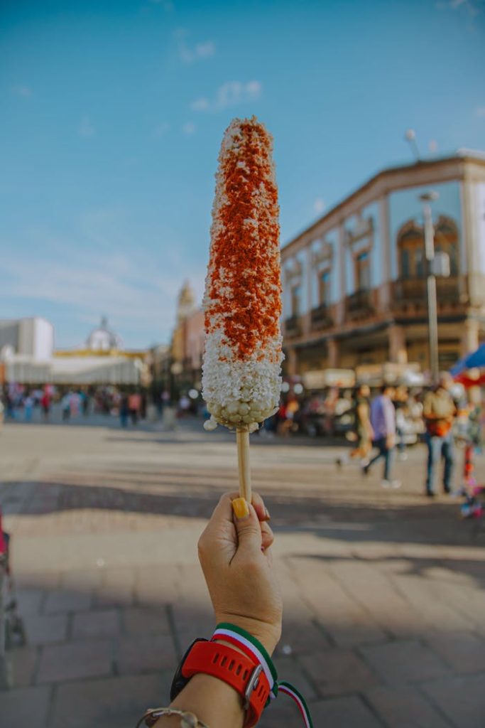 Hand holding a delicious Mexican street corn (elote) in a vibrant Irapuato market setting.