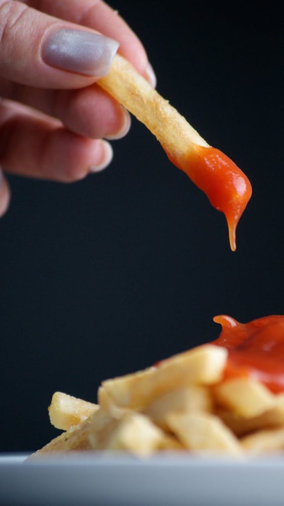 Close-up of a hand dipping a French fry in ketchup, highlighting fast food indulgence.