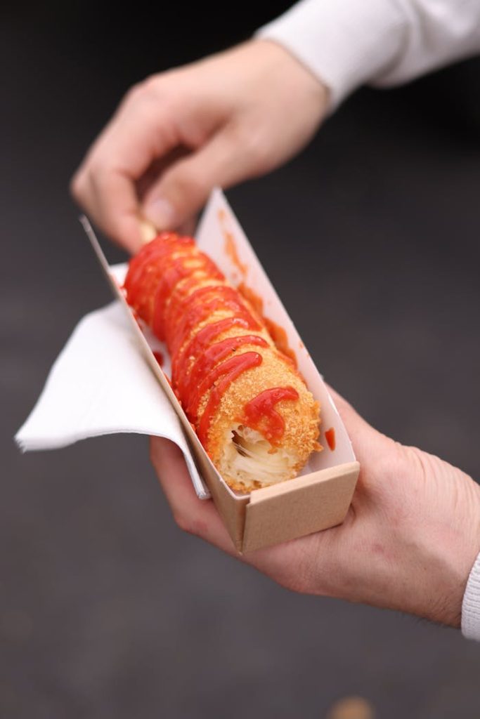 Close-up of a fried corn dog topped with ketchup held in hands, perfect street food snack.