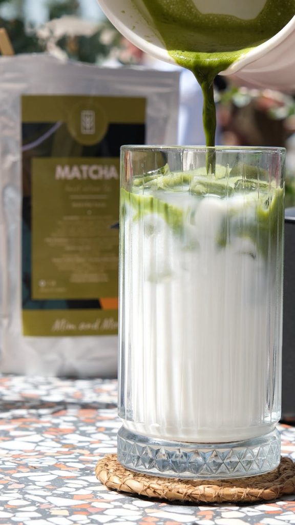 A creamy iced matcha latte being poured on a sunny day with a matcha package in the background.