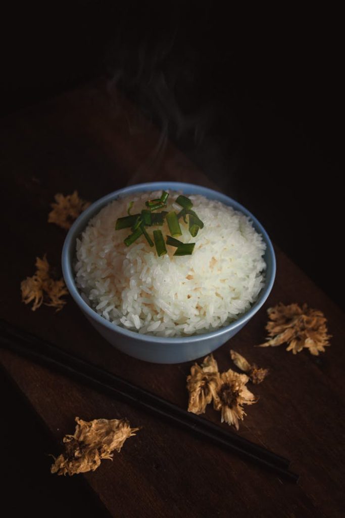 A steaming bowl of rice garnished with green onions, surrounded by dried flowers, captured in a rustic style.
